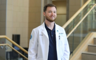 A Doctor of Osteopathy from Kansas College of Osteopathic Medicine wears a lab coat and smiles while looking off-camera.