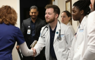 Doctor in white coat shakes hands with one in blue as others look on.