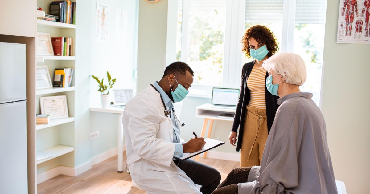 A doctor sits and writes notes while in conversation with an elderly patient and the patient's companion.