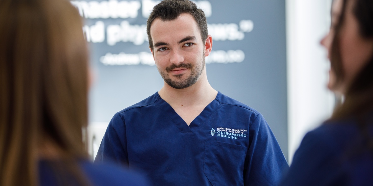 A student doctor wearing KansasCOM-branded scrubs faces two figures in the foreground with their backs to the camera.