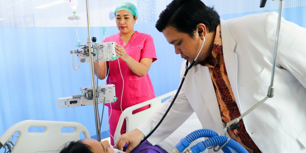 A doctor examines a patient while a nurse checks IV>